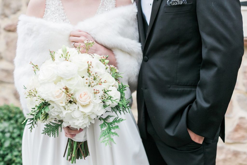 Bride wearing white fur wrap holding white floral bouquet with groom in black tux by the Jepsons