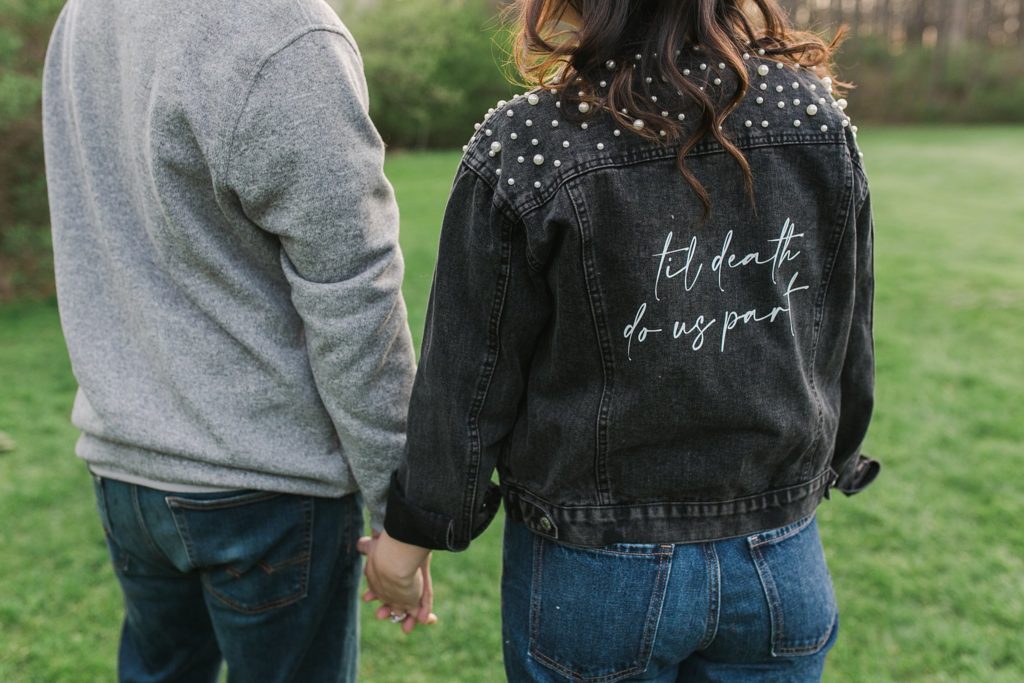 Engaged couple holding hands while bride wears jacket displaying "til death do us part" by the Jepsons